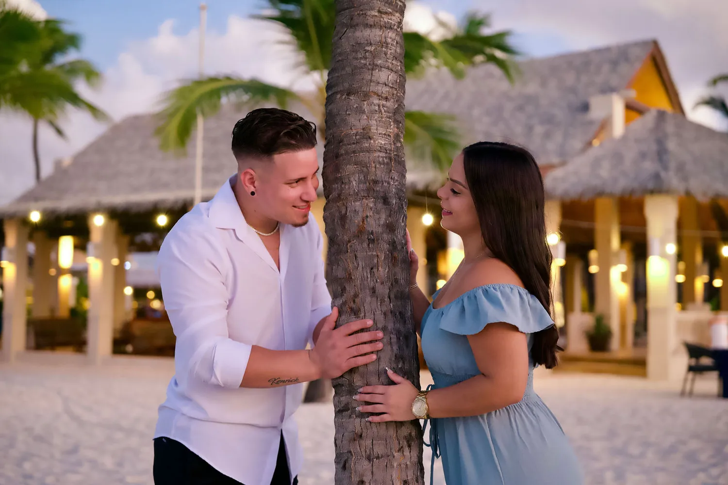 Couple leaning against a palm tree at a beach bar during their photoshoot in Aruba