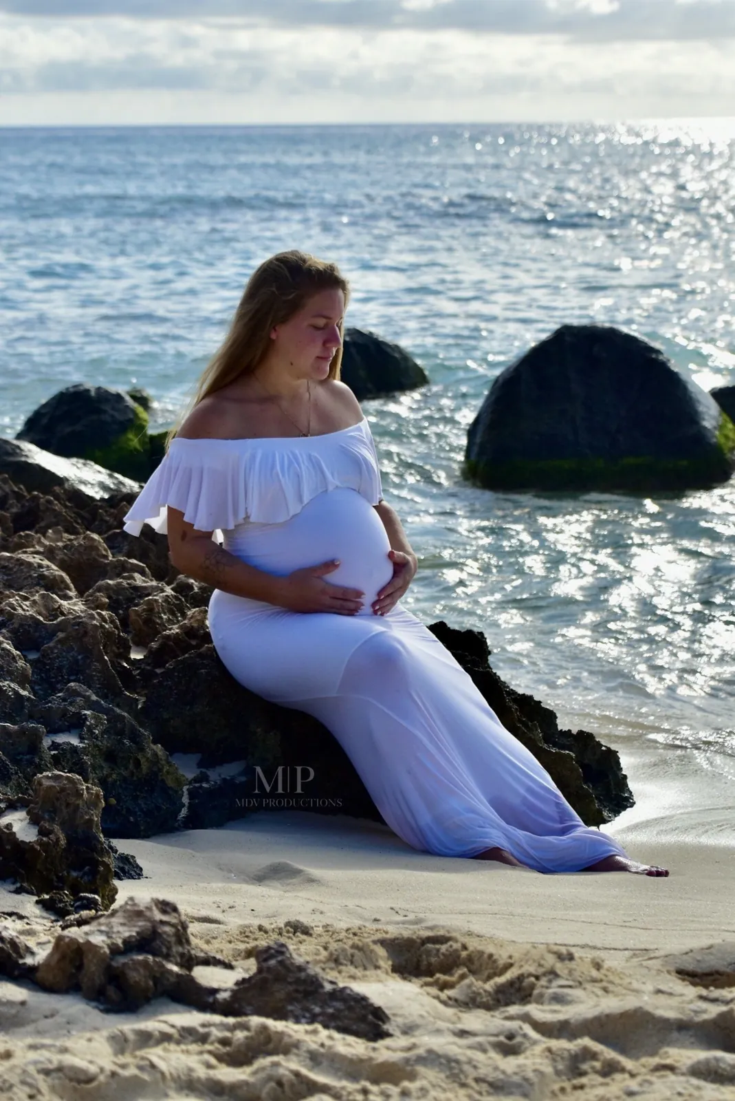 Pregnant woman in a white dress sitting on the rocks by the sea in Aruba