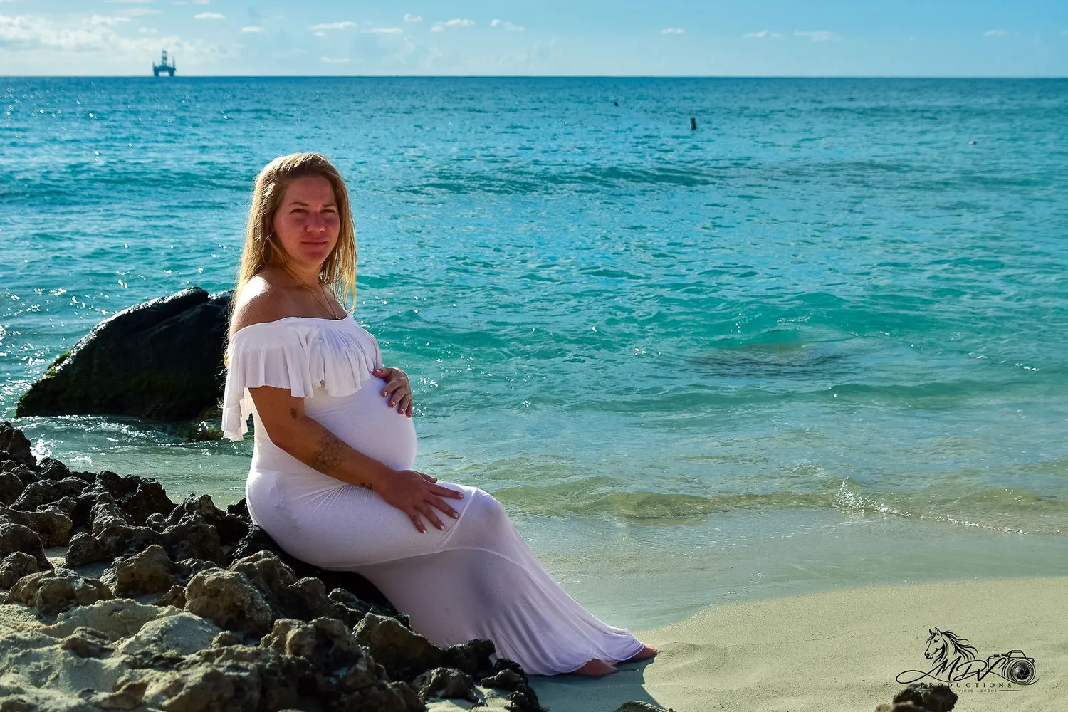 Pregnant woman in white sitting on the rocks with the sea behind her in Aruba