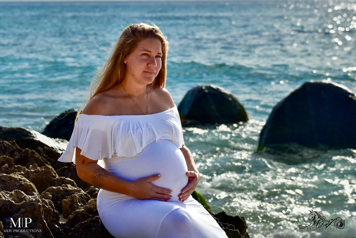 Pregnant woman in a white off shoulder dress holding her belly by the waves in Aruba